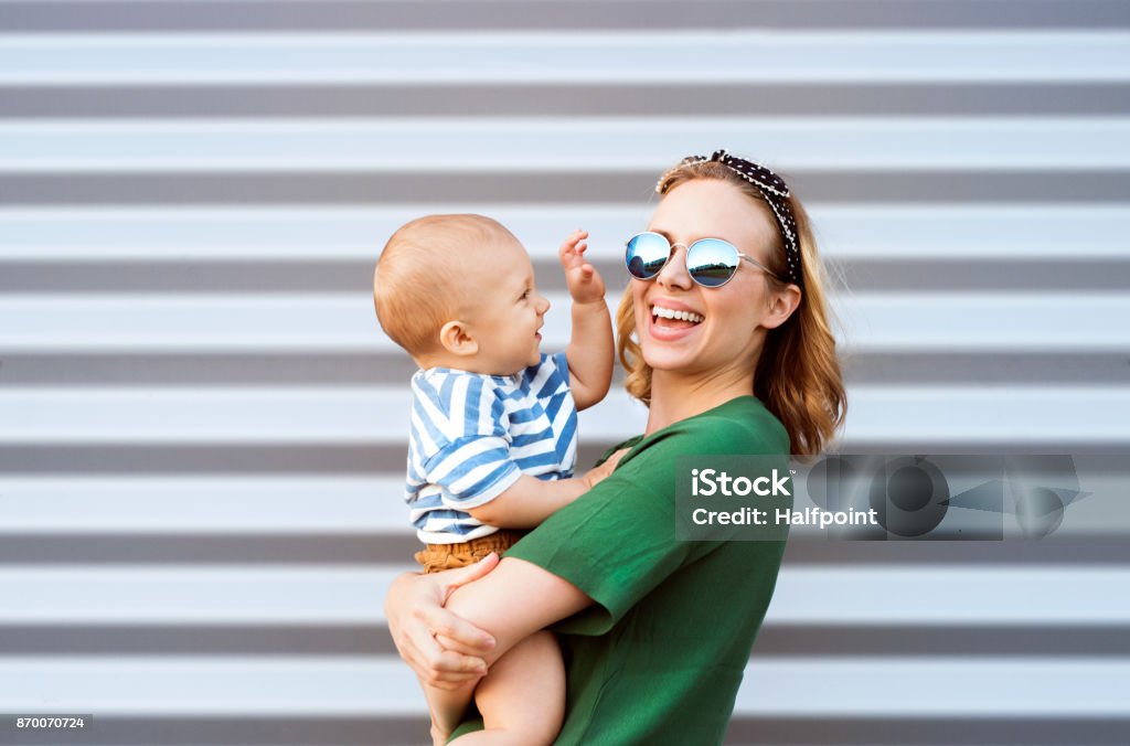 Young woman with a baby boy standing against the wall. Young woman with a baby boy standing against the wall. Beautiful mother holding her son in arms. Mother Stock Photo