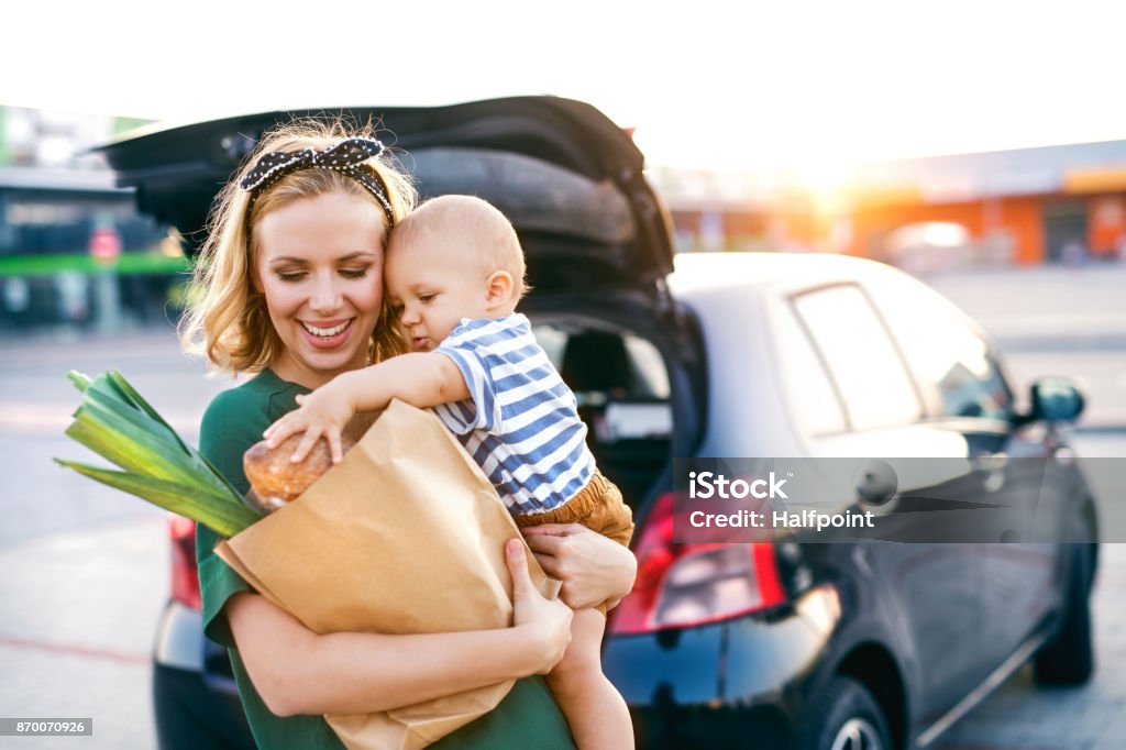 Young mother with baby boy in front of a supermarket. Beautiful young mother with her little baby son in front of a supermarket, holding paper shopping bag. Woman with a boy standing by the car. Shopping Stock Photo