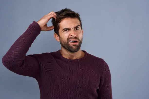 Studio shot of a young man scratching his head in confusion against a gray background
