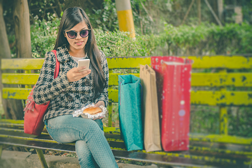 Outdoor image of beautiful Asian young woman taking break from shopping and using smart phone while eating fast food. One person and selective focus with copy space.