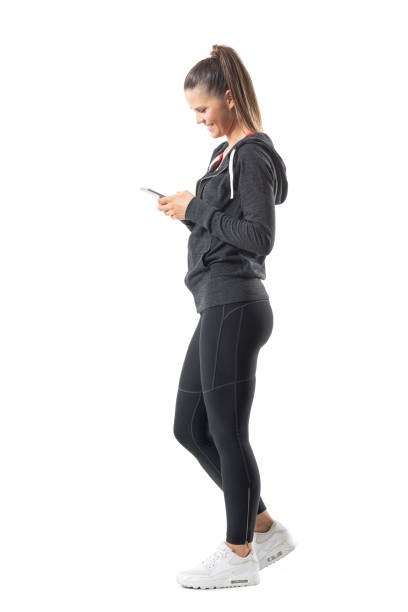 Side view of young happy fit runner woman using mobile phone and smiling Side view of young happy fit runner woman using mobile phone and smiling. Full body length portrait isolated on white background. yoga pants photos stock pictures, royalty-free photos & images