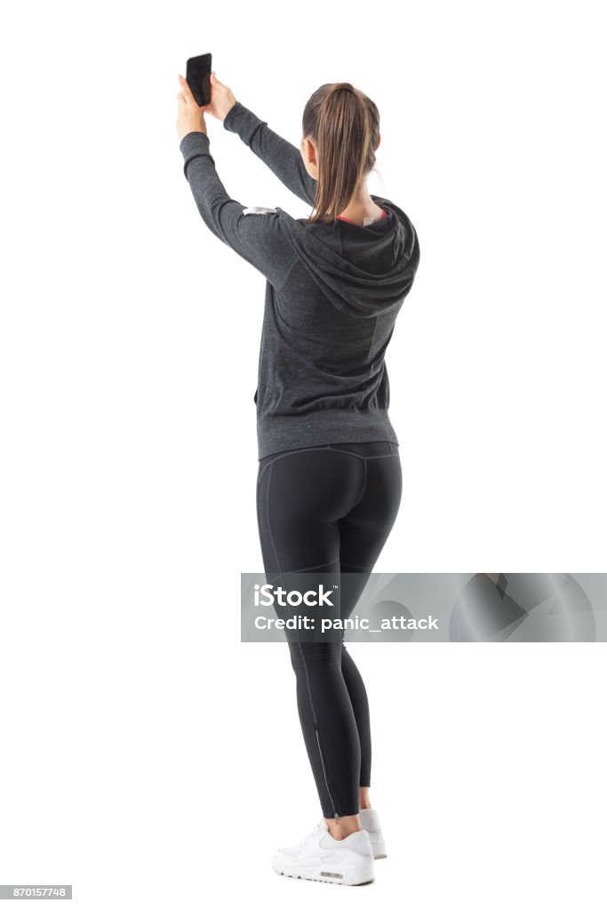 Rear view of sporty athletic woman taking selfie while holding cellphone in both hands. Rear view of sporty athletic woman taking selfie while holding cellphone in both hands. Full body length portrait isolated on white background. Full Length Stock Photo