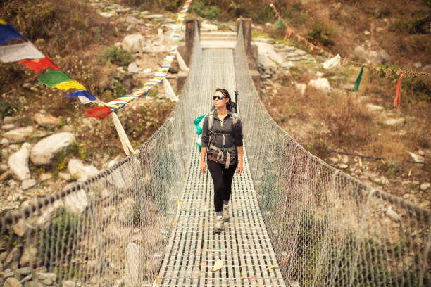 Young backpacker woman traveling in Nepal Himalayas in Annapurna conservation area and crossing over hanging bridge on mountain river. She is wearing hiking boots, black pants and green backpack.