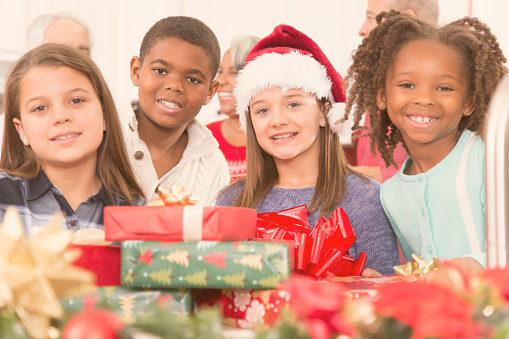 Multi-ethnic group of family members including children gather in a home kitchen to celebrate their family Christmas party and gift exchange.  Christmas tree, holiday food items, and various decorations in scene.  Cute group of children pose for camera.