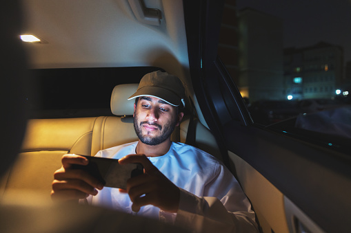 Rich young middle eastern man watching football highlights while seated in the backseat of a luxury car at night.