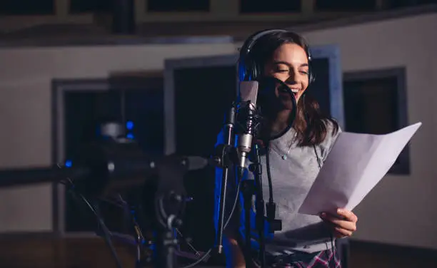Smiling female singer with microphone and reading lyrics. Woman recording a song in music studio. Female playback singer recording her new album.