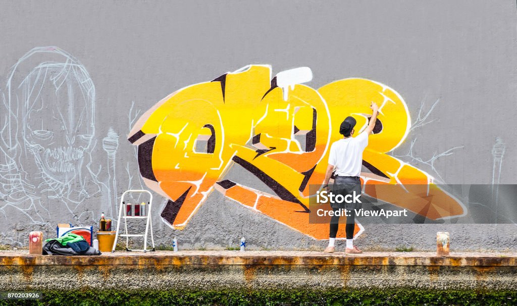 Street artist painting colored graffiti on public space wall - Modern art concept of urban guy performing and preparing live murales paint with yellow aerosol color spray - Cloudy afternoon filter Graffiti Stock Photo