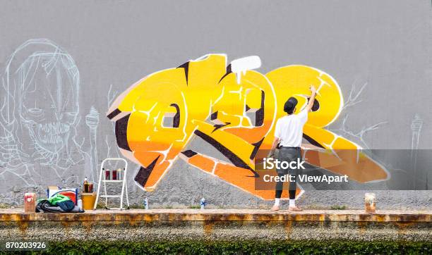 Street Artist Painting Colored Graffiti On Public Space Wall Modern Art Concept Of Urban Guy Performing And Preparing Live Murales Paint With Yellow Aerosol Color Spray Cloudy Afternoon Filter Stock Photo - Download Image Now