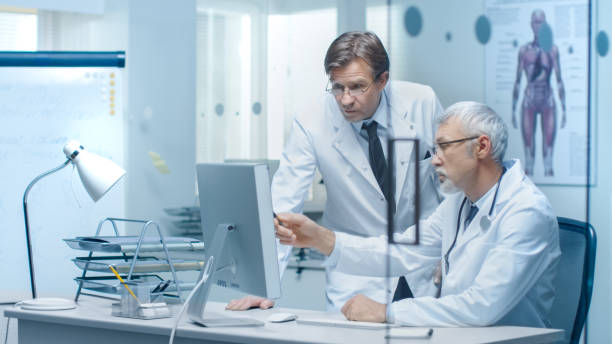 istock Senior Doctor and His Assistant Discuss Patient's Log on Personal Computer. 870233550