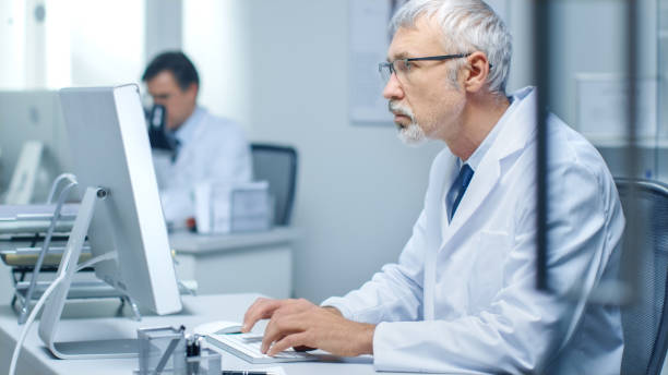 istock Senior Medical Practitioner Working in His Office at His Desktop Computer. Also His Assistant Works in the Background. 870233580