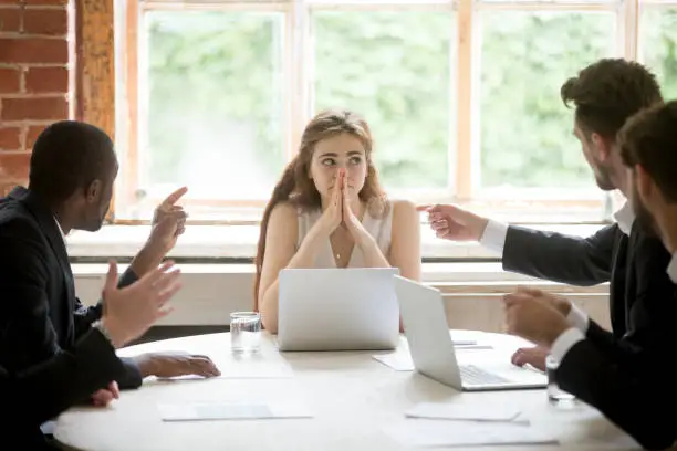 Perplexed young woman looking at coworkers pointing fingers at her. Disgruntled multiethnic team scolding female colleague who made bad work-related mistake. Stressed upset business lady in trouble.