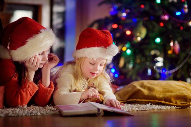 istock Two cute little sisters reading a story book together under a Christmas tree 870258614
