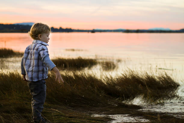 Little boy with origami boat sits on the old wooden bridge at the river on the sunset background. A child near the river looks a beautiful sunset in summer.