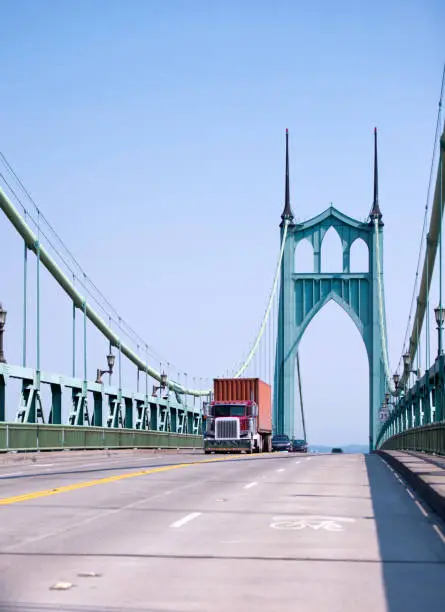 Photo of Red big rig semi truck carry container on flat bed trailer and moving on gothic arched St Johns bridge in Portland