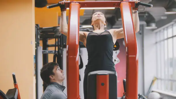 Fitness-club - young woman performs Pull-Ups with male coach, telephoto view