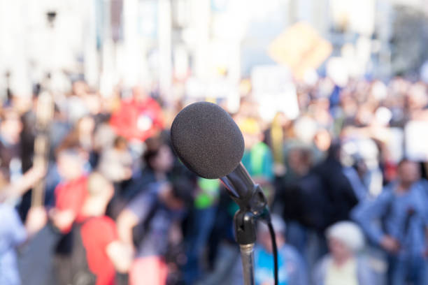 istock Protest. Public demonstration. Microphone in focus against blurred crowd. 870430550