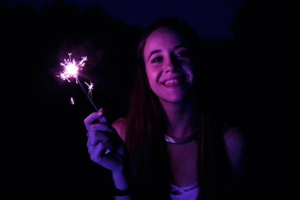 Pretty young woman standing in park at night, holding sparkler and smiling.