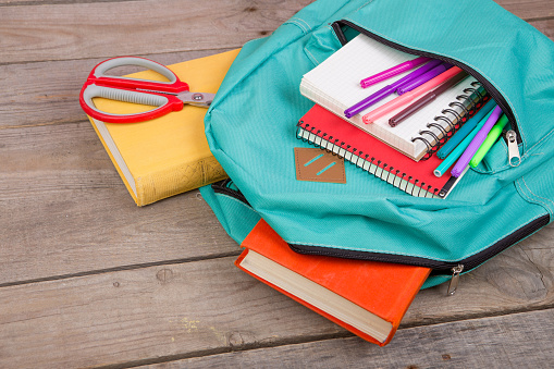 Backpack and school supplies: books, notepad, felt-tip pens, scissors on brown wooden table