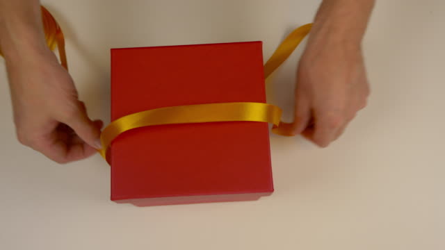 Preparation for gift wrapping. A red box of cardboard. Mens hands measure the gold satin ribbon to decorate the gift box. Top view close up. Yellow tape.