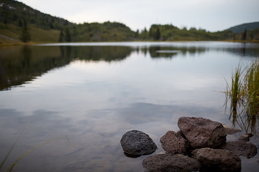 Rocks on the shoreline of a tranquil lake on a gloomy overcast day surrounded by forested hills and mountains reflected in the water
