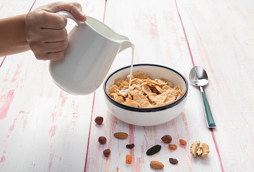 Pouring milk on a bowl of cornflakes  , hazelnut, dry apricot, walnut and nuts on wooden background , top view,  close-up