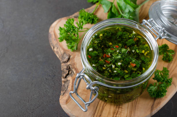 Traditional argentinian chimichurri sauce made of parsley, cilantro, garlic and chili pepper in a glass jar. Selective focus, horizontal image A glass jar wirh homemade fresh chimichurri sauce standing on a wooden board with herbs on a background chimichurri stock pictures, royalty-free photos & images