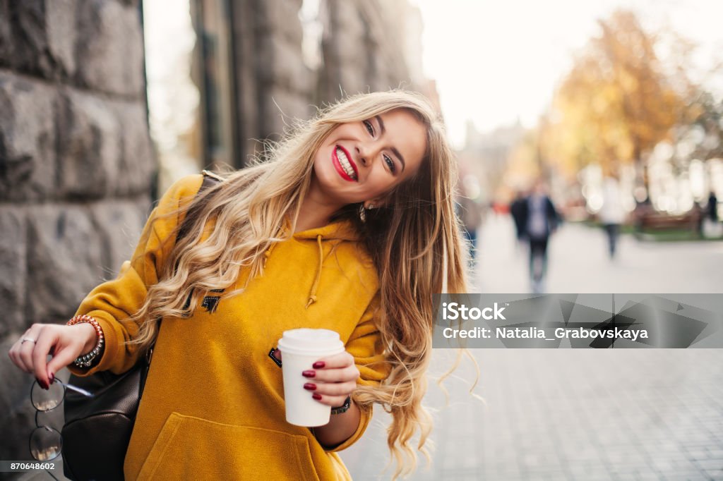 Élégante heureuse jeune femme porte un jeans boyfrend, baskets blanc lumineux jaune sweetshot. Elle est titulaire de café pour aller. Portrait de jeune fille souriante dans lunettes de soleil et avec sac - Photo de Femmes libre de droits