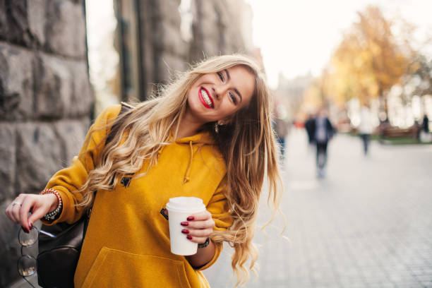세련 된 행복 한 젊은 여자 boyfrend 청바지, 흰 운동 화 밝은 노란색 sweetshot를 입고. 그녀는 서 커피를 보유 하고있다. 선글라스와 가방으로 웃는 여자의 초상화 - sensuality blond hair women beauty 뉴스 사진 이미지