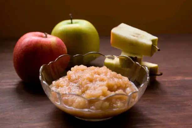 A bowl of applesauce with one red and one green whole apples and three cored apples sitting on a wood table.