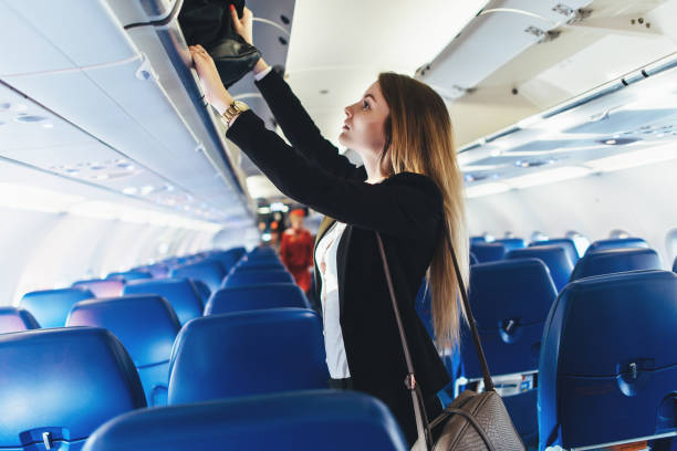 istock Female student putting her hand luggage into overhead locker on airplane 870704458