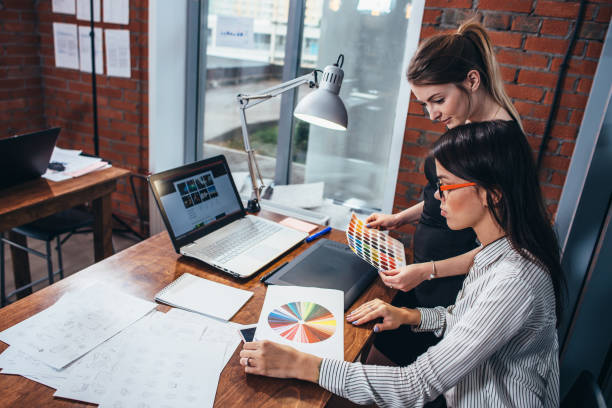 mulheres jovens trabalhando em um novo design de web usando amostras de cores e desenhos, sentado na mesa de escritório moderna - business computer monitor paper digital tablet - fotografias e filmes do acervo