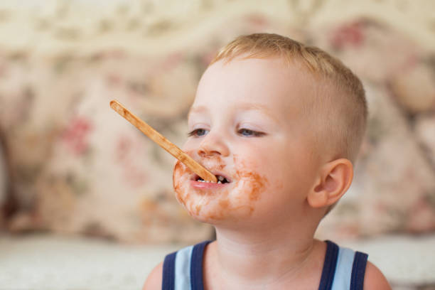 Closeup portrait of little dirty-faced boy holding ice-cream stick in his mouth and biting it