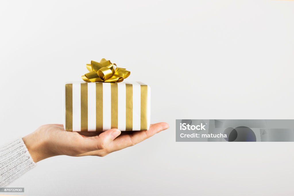 Woman's hand holding a gift in white and gold paper on a gray background. Copy space Gift Stock Photo