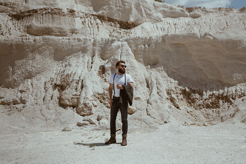 Young bearded stylish traveler posing with jacket in hands looking away on background of rocks.