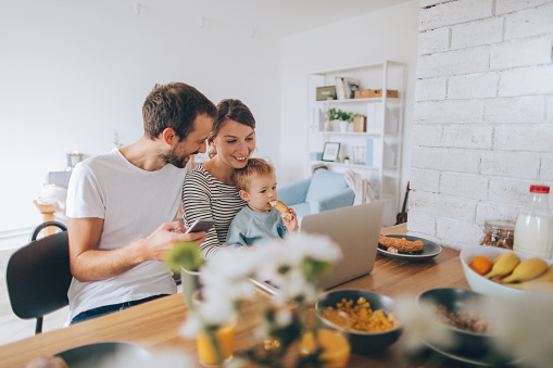 Photo of a young family is showing their hectic lifestyle - busy morning they are spending together over dinning table, using computer and mobile phone while their toddler boy is with them