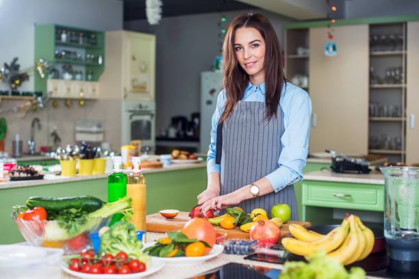 istock Young woman standing in her kitchen wearing apron cooking, cutting fruit on a board 870799458