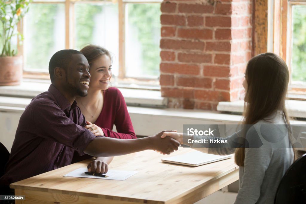 Young multiethnic couple shaking hands with real estate agent. Happy smiling african american man shaking hands with real estate agent. Husband and wife signed rental lease agreement. Young multiethnic couple got great deal, rented or purchased new home concept. Apartment Stock Photo