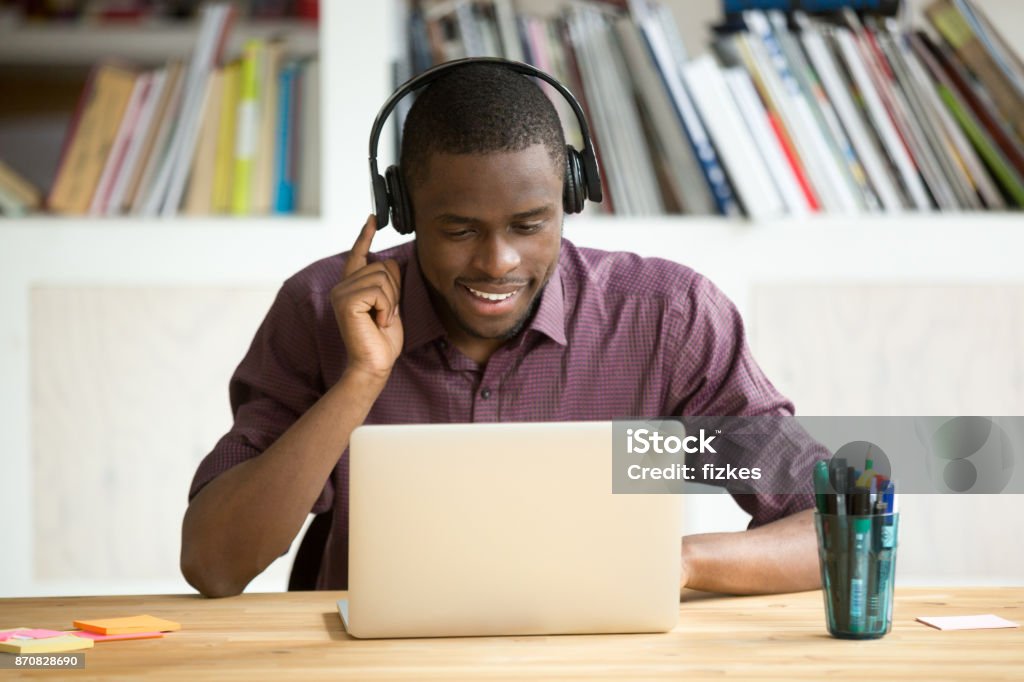 Casual smiling office worker in headphones looking at laptop screen. Smiling african american office worker in headphones looking at laptop screen. Young  casual businessman studying foreign language, communicating with clients through video conference application. Media Interview Stock Photo