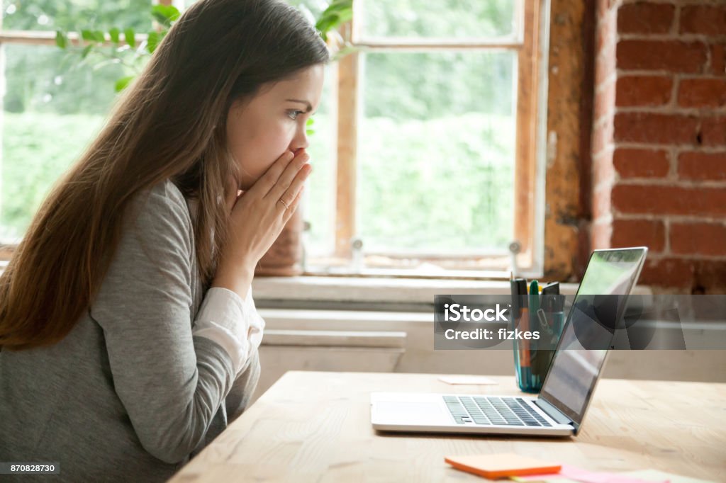 Shocked young woman looking at laptop screen at work desk. Shocked young woman looking at laptop screen at work desk. Casual business lady in office seeing something unbelievable on computer. Bad news concept, dismissal notice from boss, work-related mistake. Shock Stock Photo