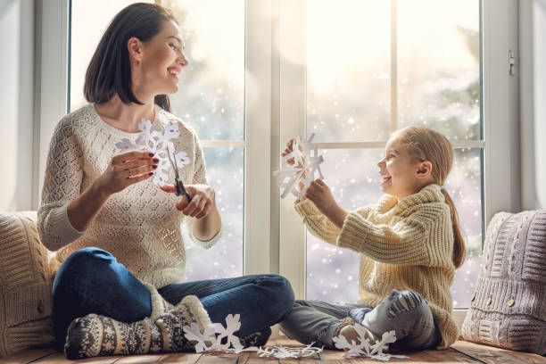 family sitting by the window Merry Christmas and happy holidays! Happy loving family sitting by the window and making paper snowflakes for decoration windows. Mother and child creating decorations. children in winter stock pictures, royalty-free photos & images