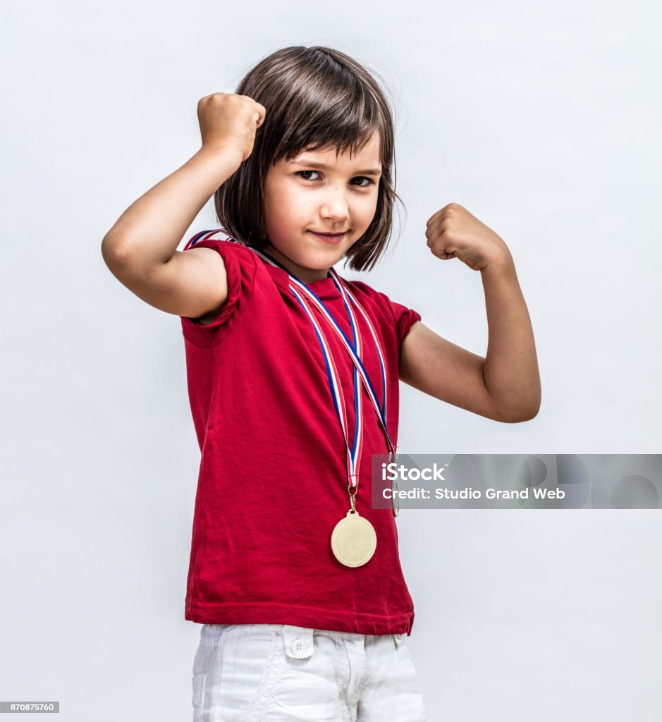 Little cheeky girl with medals smiling, showing young female power Little cheeky girl with medals smiling with strong arms raised, showing her young female power, motivation and winning success over a light grey background Child Stock Photo