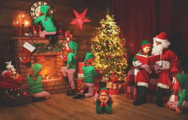Santa Claus and little elves before Christmas in his house by fireplace and Christmas tree