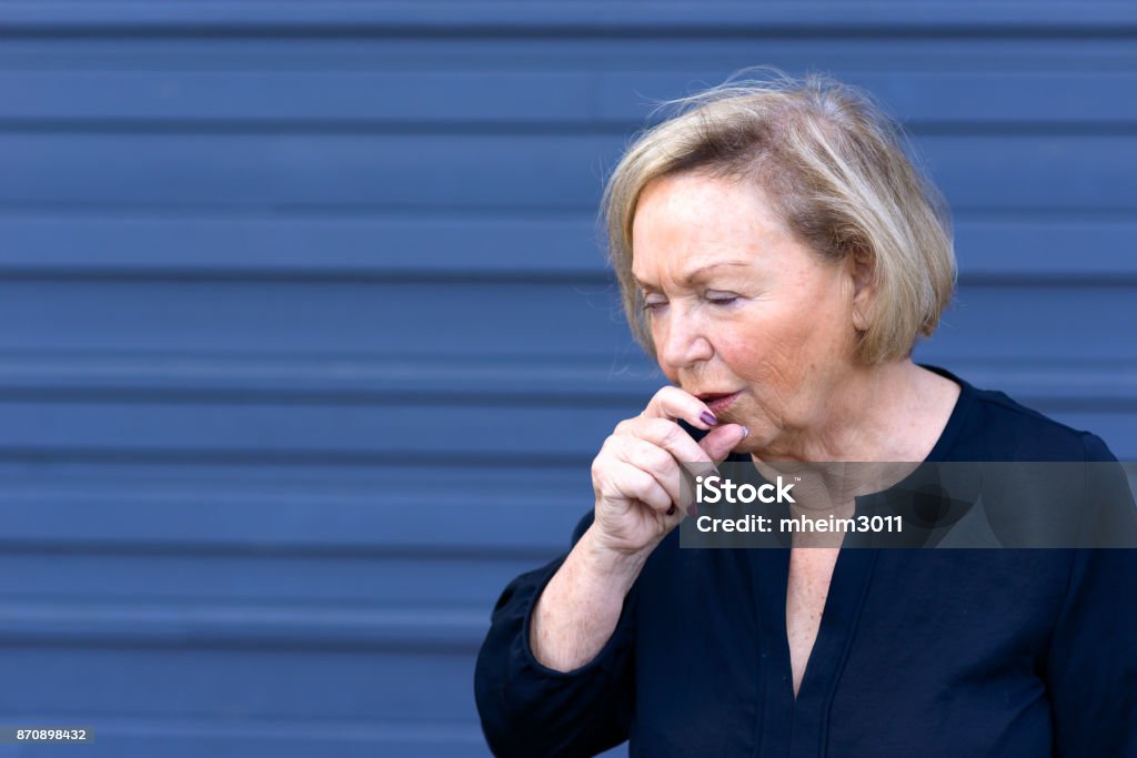 Unwell elderly lady having a coughing fit Unwell elderly lady having a coughing fit holding her hand to her mouth conceptual of seasonal flu, allergies or choking over blue with copy space Choking Stock Photo