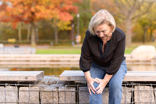 Elderly lady grabbing her knee in pain as she sits on a wall surrounding a pond after injuring herself out walking on an autumn day with copy space