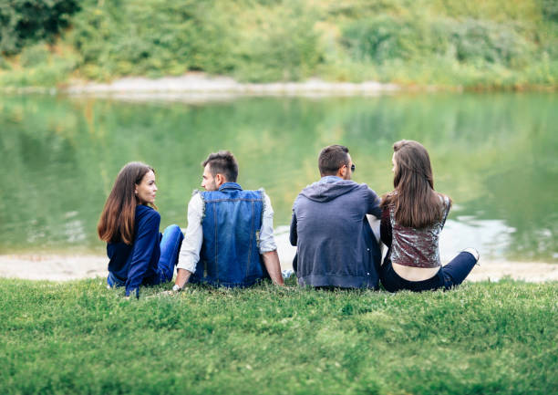 istock Friends sitting on grass by lake in park back view 870923196