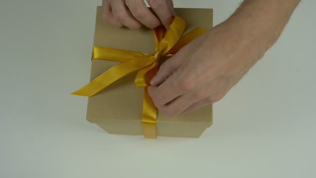 Close up top view high angle. Mens hands straighten a gold bow from a satin ribbon on a gift box. Preparing for the gift giving event. Gift box with yellow bow. Brown carton box.