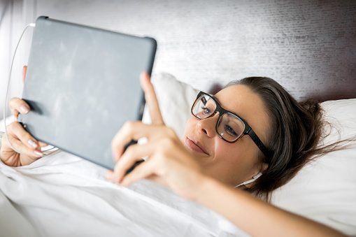 Beautiful Young Woman Using digital tablet in Bed at home or in hotel room. She is looking at the screen with headphones.
