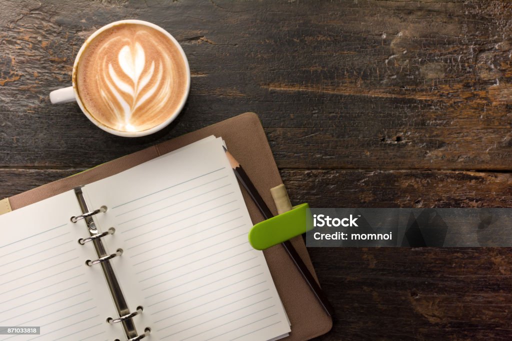 Open notebook with a cup of latte coffee on table. A cup of coffee with latte art tulip on top next to blank note book with pencil on old wood background, top view. Open notebook with a cup of latte coffee on table. Note Pad Stock Photo