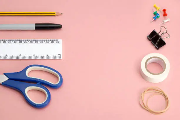 Office stationery and other supplies including a pair of scissors, ruler, marker pen, a pencil, thumbtacks, a binder clip, paper adhesive tape, and rubber bands on a pink background with copy space
