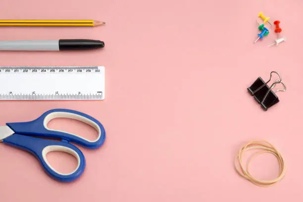 Various office or school supplies and tools including a pair of scissors, ruler, marker pen, a pencil, push pins, a binder clip and rubber band on a pink background with copy space
